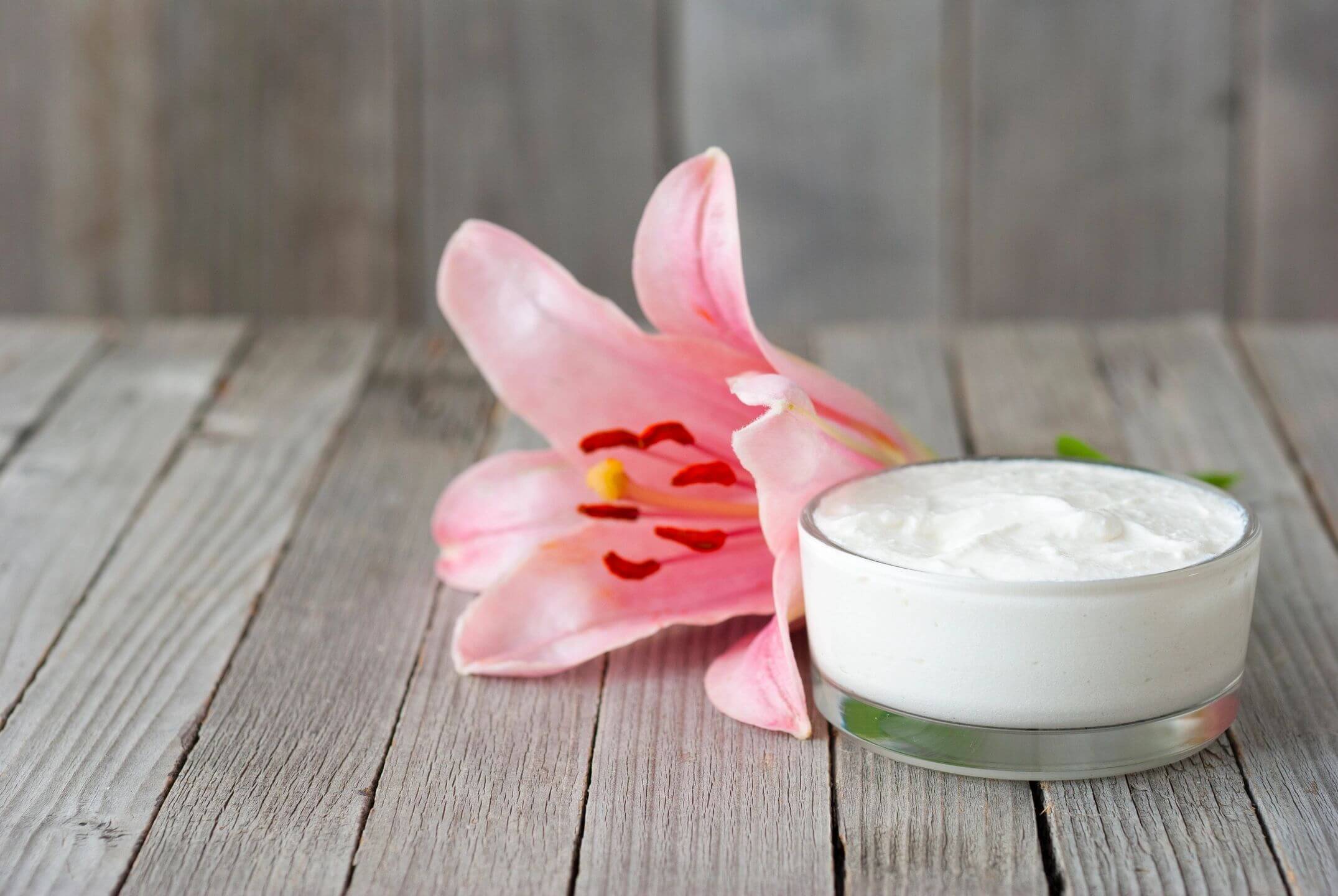 Pink lily flower beside a container of white cream on wooden surface.